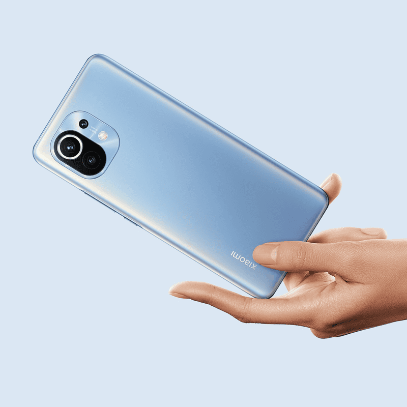 Smartphone Xiaomi Mi 11 yang sedang dipegang menampilkan bagian belakang dan terlihat ada triple camera dari smartphone. Smartphone Xiaomi Mi 11 berwarna biru muda.